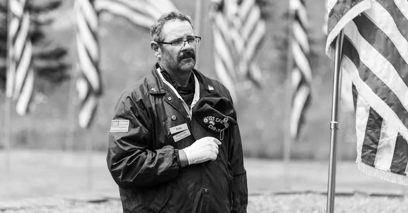 A veteran holding a hat stands solemnly among flags at a military memorial in black and white.
