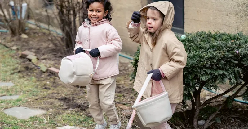 Two children happily engage in an Easter egg hunt wearing winter coats outside a building.