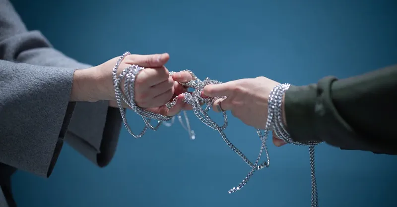 Close-up of two hands wrapped in silver chains, symbolizing connection or struggle, set against a blue backdrop.