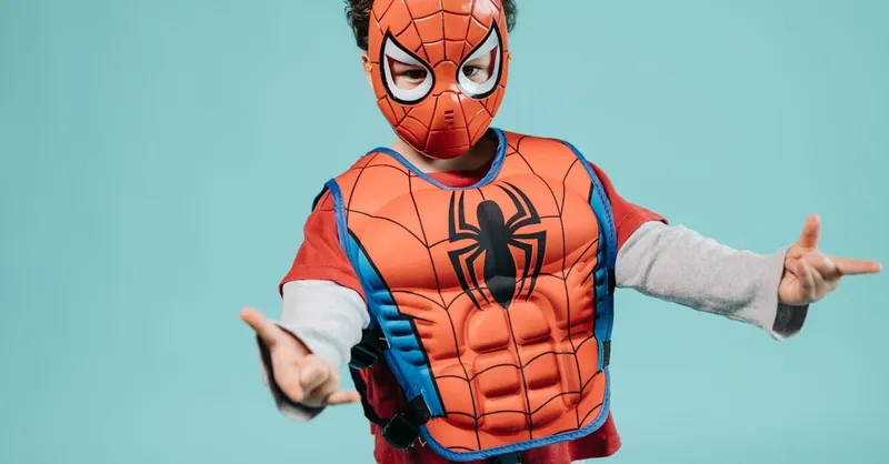 Child wearing a Spiderman costume and mask striking a playful superhero pose in a studio setting.