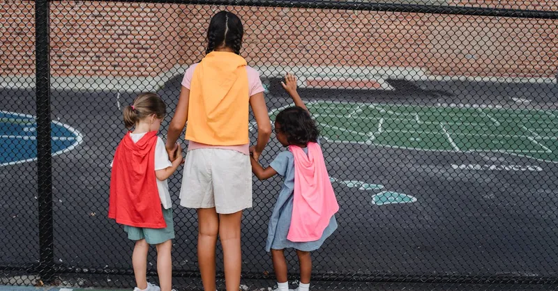 Three kids dressed as superheroes peer through a playground fence, embracing diversity and imagination.